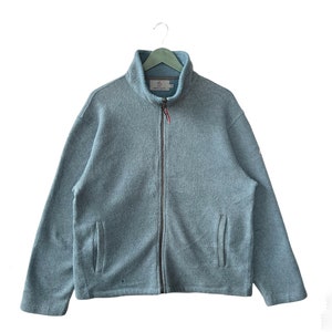 Puede incluir: Chaqueta polar azul claro con cuello alto y cremallera completa. La chaqueta tiene dos bolsillos delanteros y un pequeño logotipo en la manga. Cuelga de una percha verde.