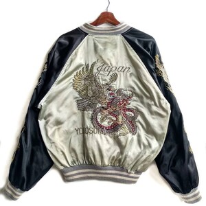 May include: A black and silver satin bomber jacket with an embroidered eagle and dragon design on the back. The jacket has a ribbed collar, cuffs, and waistband. The text "Japan" and "Yokosuka" are embroidered on the back.