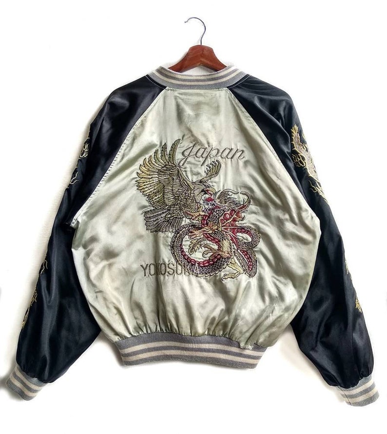 May include: A black and silver satin bomber jacket with an embroidered eagle and dragon design on the back. The jacket has a ribbed collar, cuffs, and waistband. The text "Japan" and "Yokosuka" are embroidered on the back.