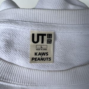 以下が含まれることがあります： UTのロゴ、サイズL、前面に「KAWS x PEANUTS」の文字がプリントされた白い長袖スウェットシャツです。