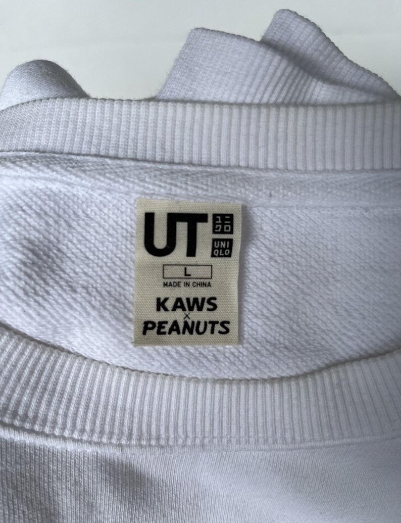 以下が含まれることがあります： UTのロゴ、サイズL、前面に「KAWS x PEANUTS」の文字がプリントされた白い長袖スウェットシャツです。