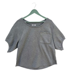 May include: A gray short-sleeved t-shirt with a pocket on the left side. The shirt has a round neckline and is hanging on a light green hanger. The label on the shirt reads "Acne".