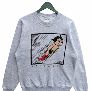 May include: A gray sweatshirt with a graphic print of Astro Boy flying. The print is in a black frame with the text "ASTRO BOY." printed below the image.