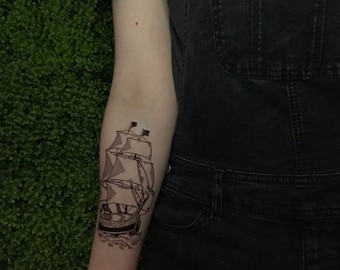 May include: A black and white line drawing of a sailing ship with three sails, a mast, and a rudder. The ship is on a wave and has a flag on the top of the mast. The drawing is on a person's arm.