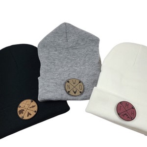 Op de afbeelding: Drie gebreide mutsen in zwart, grijs en wit met verschillende ronde houten patches. De zwarte muts heeft een patch met een beer, een vis en pijlen. De grijze muts heeft een patch met de letters NWO en een hertenkop. De witte muts heeft een patch met de letters IMH en een berg.