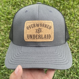 May include: A grey and black trucker hat with a cork patch that reads "Overworked and Underpaid".