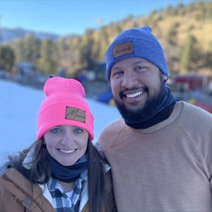 Puede incluir: Dos gorros de punto, uno rosa brillante con un parche "Mama" y otro azul con un parche "Twerdi". Los gorros se llevan al aire libre en un entorno nevado.