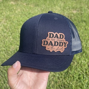 May include: A black and navy blue trucker hat with a brown leather patch that reads "Dad in the streets Daddy in the sheets".