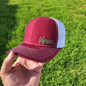 以下が含まれることがあります： 前面に茶色の金属製の「ママ」パッチが付いた、マルーンと白のトラッカーハットです。