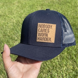 Puede incluir: Una gorra de camionero negra y azul marino con un parche de corcho que dice "Nobody Cares Work Harder."