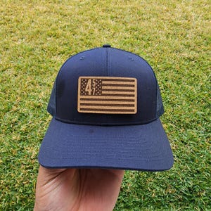 Op de afbeelding: Een navy blauwe trucker cap met een mesh achterkant. De pet heeft een kurk patch met een Amerikaanse vlag en de nummers "457" erop gedrukt.
