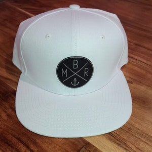 Op de afbeelding: Witte baseballpet met een zwart cirkelvormig logo met de letters "BMR" met een kruis en een anker erin.
