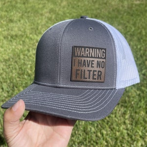 Peut inclure: Un chapeau de camionneur gris et blanc avec un patch en cuir marron qui dit "WARNING I HAVE NO FILTER".