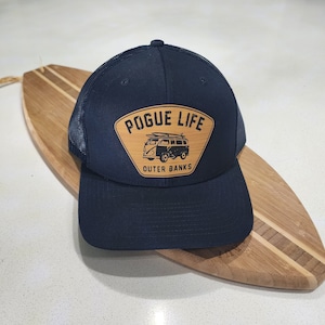 May include: A black trucker hat with a brown patch that says "Pogue Life Outer Banks" and features a black and white illustration of a van with surfboards on top.