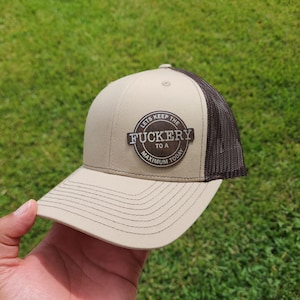 以下が含まれることがあります： ベージュとブラウンのトラッカーハット。ブラウンの革のパッチには「Let's Keep The Fuckery To A Maximum Today」と書いてあります。