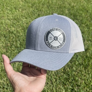 Op de afbeelding: Een grijze en witte truckercap met een zwart-witte patch waarop "Sore and Back for More" staat met een gewichtsplaatontwerp. De gewichtsplaat geeft "45 LBS" en "20.4 KGS" aan.