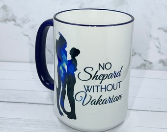 May include: White ceramic mug with a navy blue handle and rim. The mug features a silhouette of two figures with blue watercolor accents and the text "NO Shepard WITHOUT Vakarian".