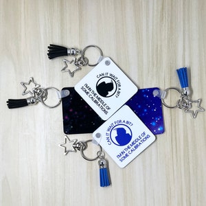 May include: Three white square keychains with blue and black accents. Each keychain features a blue and white star charm, a tassel, and a silver ring. The keychains have the text "Can it wait for a bit? I'm in the middle of some calibrations." printed on them.