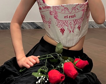 May include: A white corset with a red floral pattern and the text "no end" on a white banner. The corset is worn over a black skirt and the person is holding a bouquet of red roses.