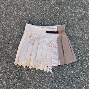 Puede incluir: Una falda corta con un diseño de patchwork. Un lado es una falda blanca con ribete de encaje y el otro lado es una falda a cuadros con un patrón marrón, rojo y blanco.