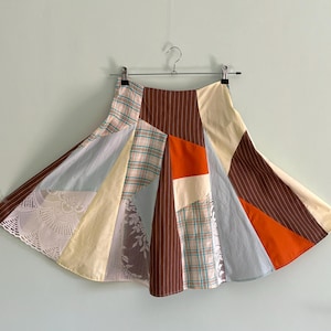 Op de afbeelding: Een patchwork rok met een verscheidenheid aan stoffen, waaronder ruitjes, strepen en kant. De rok heeft een mix van kleuren zoals bruin, oranje, crème en lichtblauw. Hij hangt aan een metalen hanger tegen een lichtgroene achtergrond.