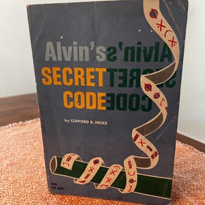 May include: A vintage book titled "Alvin's Secret Code" by Clifford B. Hicks. The cover is dark blue with orange and green lettering and a decorative ribbon design. The book is displayed on an orange textured surface.