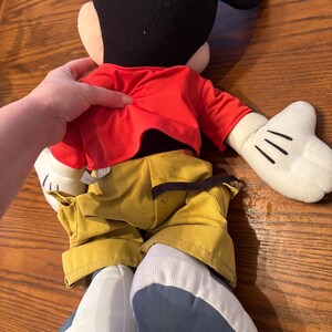 May include: A large Mickey Mouse plush toy, viewed from the back. The plush has a black head with large ears, a red jacket, yellow pants, and white gloves. The shoes are blue and white. The toy is on a wooden surface.