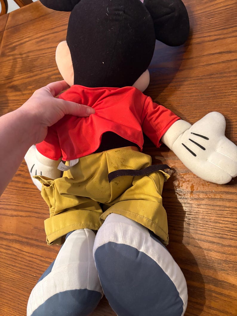 May include: A large Mickey Mouse plush toy, viewed from the back. The plush has a black head with large ears, a red jacket, yellow pants, and white gloves. The shoes are blue and white. The toy is on a wooden surface.