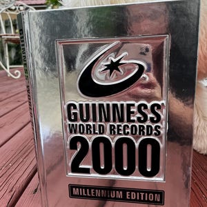 May include: A silver-colored Guinness World Records 2000 Millennium Edition book. The cover features the Guinness logo and the text "GUINNESS WORLD RECORDS 2000" in bold black letters. The book is on a wooden surface.