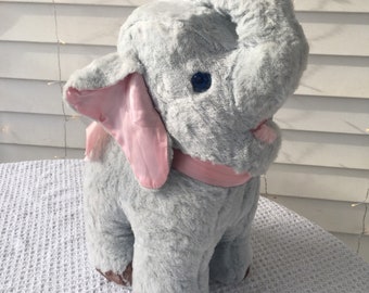 May include: A plush gray elephant toy with a raised trunk and large ears. The elephant has pink inner ears and a pink scarf. It has brown feet and blue eyes. The toy is sitting on a white surface.