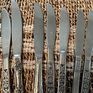 May include: A set of eight silver dinner knives with floral designs on the handles. The knives are arranged in a row on a woven straw surface.