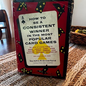 May include: A vintage book titled "How to Be a Consistent Winner in the Most Popular Card Games" is displayed. The cover is red with black card suit illustrations and text. The book details winning strategies for various card games.