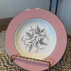 May include: A pink and white decorative plate with a floral design. The plate has a silver rim and is displayed on a gold-colored metal stand. The floral design features gray leaves and pink accents.