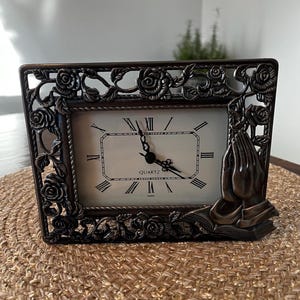 May include: A decorative, ornate, bronze-colored clock with a white face and Roman numeral markings. The clock has a floral design with roses and a praying hands motif. The clock face reads "QUARTZ" and "JAPAN".