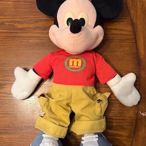 May include: A plush Mickey Mouse doll, featuring black ears, a pink face, and a red shirt with a green and yellow "mickey" logo. The doll wears yellow pants and blue and white shoes. The doll is lying on a wooden surface.