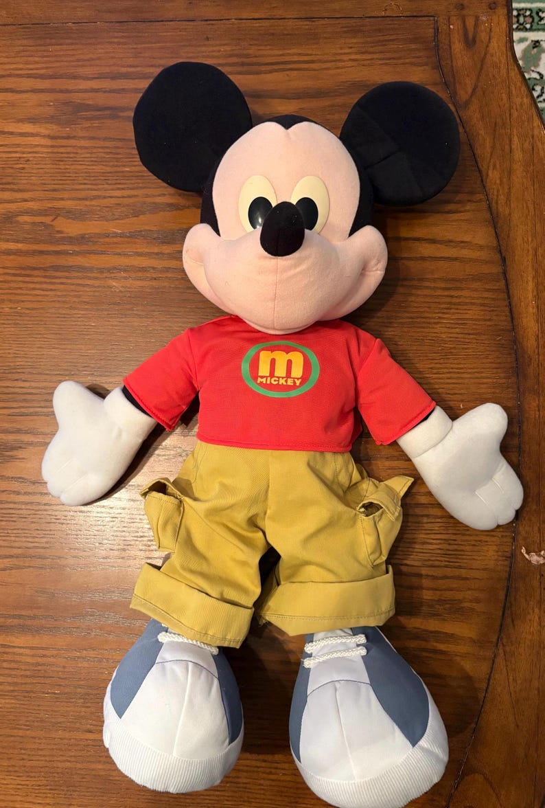 May include: A plush Mickey Mouse doll, featuring black ears, a pink face, and a red shirt with a green and yellow "mickey" logo. The doll wears yellow pants and blue and white shoes. The doll is lying on a wooden surface.