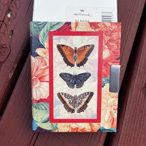 May include: A floral-patterned photo album with a red border featuring three butterfly illustrations. The album has a silver clasp and the Hallmark logo is visible. The background shows a wooden surface.