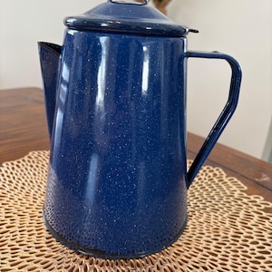 May include: A blue speckled enamel coffee pot with a lid and handle. The pot has a spout for pouring and is sitting on a woven placemat. The pot is approximately 10 inches tall.