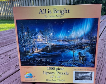 May include: A 1000 piece jigsaw puzzle titled "All is Bright" by James Meger. The puzzle depicts a winter scene with a snowy river, a cabin, and a deer in the foreground. The puzzle is 19 inches by 30 inches and has a recommended age of 10 and up.