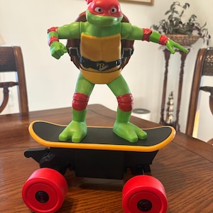 May include: A green turtle action figure with a red mask, elbow pads, and knee pads, wearing a yellow chest plate and a black belt with an R buckle. The figure stands on a black skateboard with a yellow edge and four large red wheels.