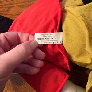 May include: A close-up of a Jumbo Mickey plush toy. The toy has a red outfit, yellow shirt, and white gloves. A tag reads "Surface Washable Only" and "1981EL". The toy is made of polyester fiber and plastic stiffener.