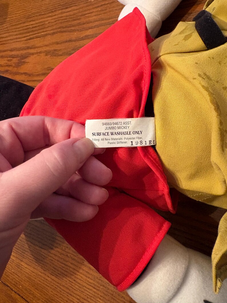 May include: A close-up of a Jumbo Mickey plush toy. The toy has a red outfit, yellow shirt, and white gloves. A tag reads "Surface Washable Only" and "1981EL". The toy is made of polyester fiber and plastic stiffener.