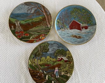 May include: Three decorative ceramic plates with raised 3D images of rural scenes. The plates depict a farmhouse in a winter scene, a farmhouse in a spring scene, and a farmhouse in a fall scene.