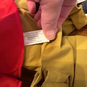 May include: Close-up of a yellow fabric garment with a red fabric item on the left. A hand is holding the yellow fabric, revealing a tag with text including "Fisher-Price, Inc." and "MADE IN CHINA."