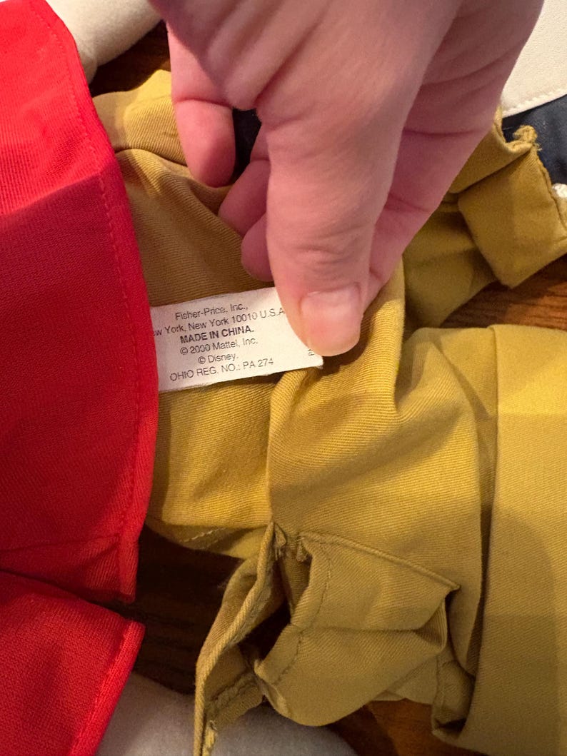 May include: Close-up of a yellow fabric garment with a red fabric item on the left. A hand is holding the yellow fabric, revealing a tag with text including "Fisher-Price, Inc." and "MADE IN CHINA."