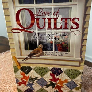 以下が含まれることがあります： 「Love of Quilts」というタイトルの本の表紙には、鳥とキルトのデザインが描かれた窓のシーンが描かれています。「A Treasury of Classic Quilting Stories」というテキストと著者の名前が記載されています。キルトのデザインには葉の模様が使用されています。