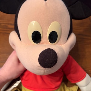 May include: A plush Mickey Mouse doll with a red shirt, yellow pants, and black ears. The doll has large, oval eyes and a black nose. The doll is being held by a person.