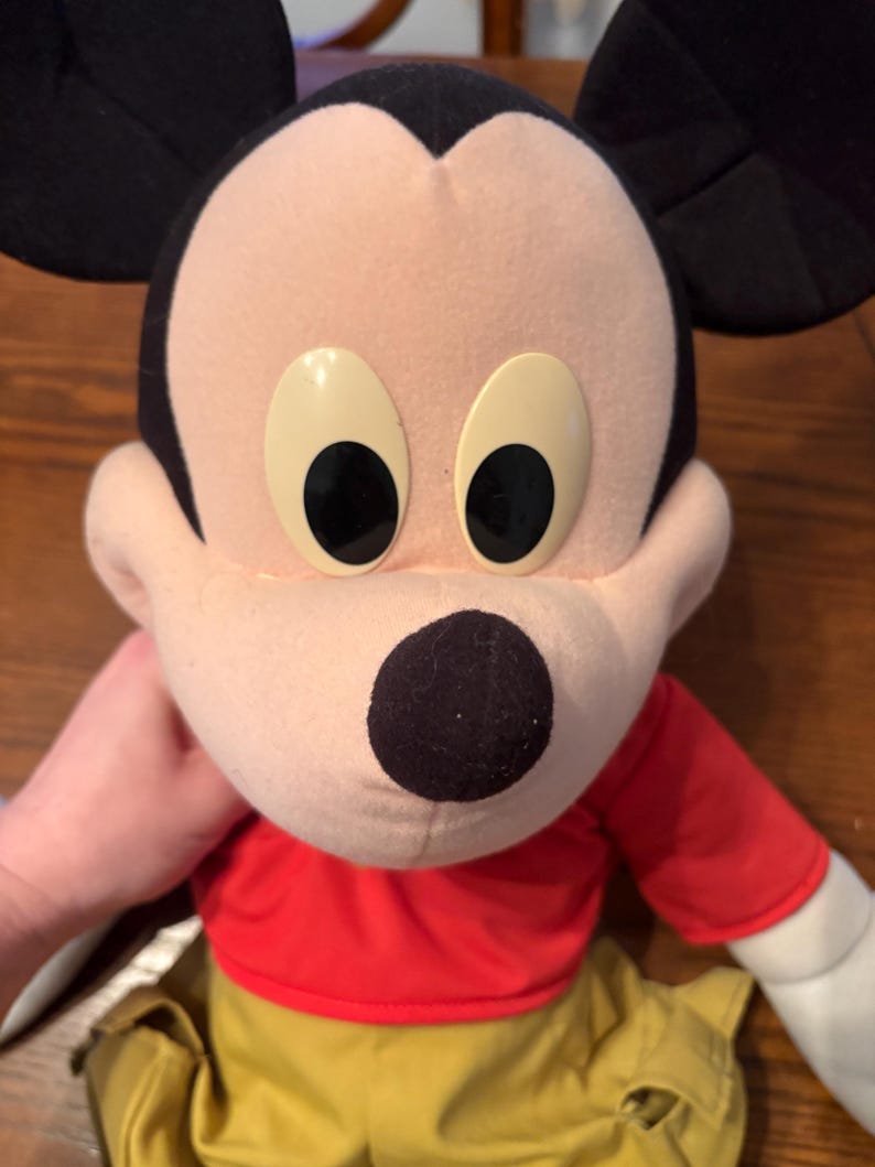 May include: A plush Mickey Mouse doll with a red shirt, yellow pants, and black ears. The doll has large, oval eyes and a black nose. The doll is being held by a person.