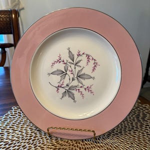 May include: A round, pink plate with a silver rim. The center of the plate is white with a floral design in gray and purple. The plate is displayed on a gold-colored metal stand.