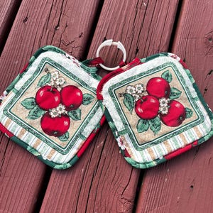 May include: Two square quilted pot holders with a red apple design. The pot holders have a white border with a green and red floral pattern. A white plastic ring connects the two pot holders.